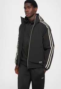 Črno puhovko s kapuco, z začimbami kremnih črt po rokavih, z zadrgo spredaj in logotipom na spodnjem robu.