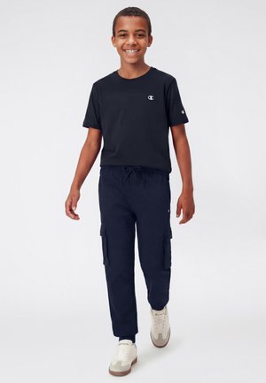 Uśmiechnięty chłopiec idący do przodu, ubrany w czarną koszulkę Champion, ciemne spodnie typu cargo oraz białe trampki Champion na jednolitym tle.