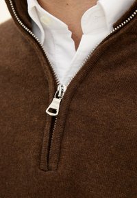 Brauner Pullover mit einem Reißverschluss-Kragen, aus weichem Material gefertigt. Auffällige Details sind ein silberner Reißverschluss und ein darunterliegendes, geschichtetes weißes Hemd.