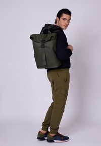 Rucsac de tip roll-top de culoare verde măslinie, cu bretele ajustabile, închidere cu cataramă și un exterior neted, rezistent la apă; detaliu cu logo pe lateral.