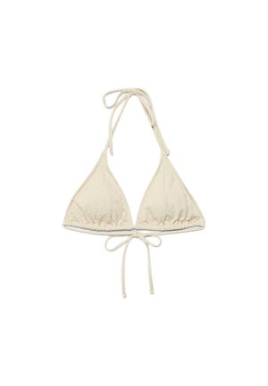 Femme aux cheveux mouillés, debout les bras croisés, portant un bikini beige et des tongs noires sur un fond marron uni.