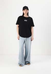 Svart bomull t-skjorte med "GAP" logo, kombinert med lyseblå vid bukse. Modellen har på seg en svart caps og brune sko.