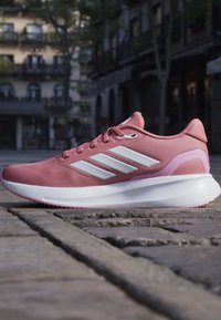 Chaussure de course rose avec des rayures blanches et une semelle blanche épaisse sur une rue pavée en milieu urbain.