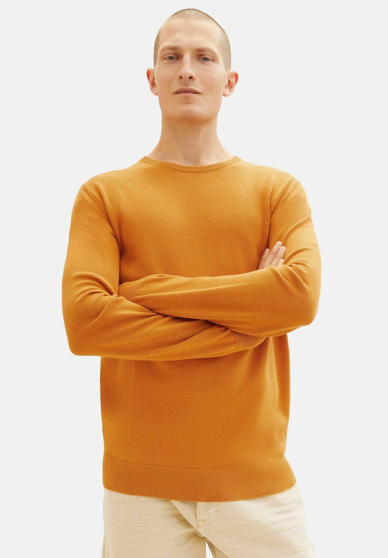 Suéter de punto naranja con escote redondo, mangas largas y puños acanalados. La modelo tiene los brazos cruzados frente a un fondo neutro.