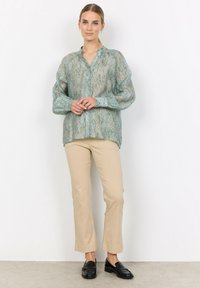 Lightweight, sheer teal blouse with patterned design, button front, and gathered collar. Paired with beige fitted pants and black loafers.