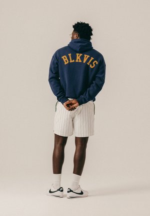 Hombre de pie con las manos entrelazadas detrás de la espalda, vistiendo una sudadera con capucha azul marino con la inscripción "BLKVIS", pantalones cortos blancos con rayas finas, calcetas blancas y zapatillas Nike blancas.