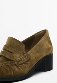 Ruskea mokkanahkainen loaferi, jossa on matala, lohkare korko; siinä on koristeellinen ommeltu nauha edessä ja hienovarainen ryppy varpaiden lähellä.