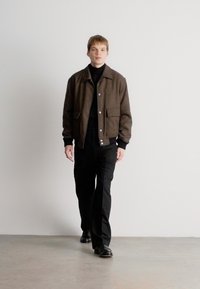 Casaco em mistura de lã castanha com fecho de botão, gola alta preta e calças pretas bem ajustadas. O modelo usa sapatos pretos. Design simples e minimalista.