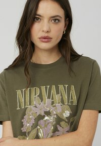 T-shirt en coton vert olive présentant le texte "NIRVANA" en blanc avec des accents métalliques dorés, embelli de graphiques floraux violets. Manches courtes.