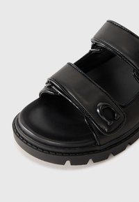 Black leather sandal with two adjustable straps, round toe, and textured rubber sole. Features a glossy finish and logo detail on the strap.