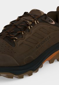 Καφέ και μαύρο παπούτσι πεζοπορίας Merrell με κορδονάκια με πορτοκαλί λεπτομέρειες και ανθεκτική σόλα για χρήση σε εξωτερικά μονοπάτια.