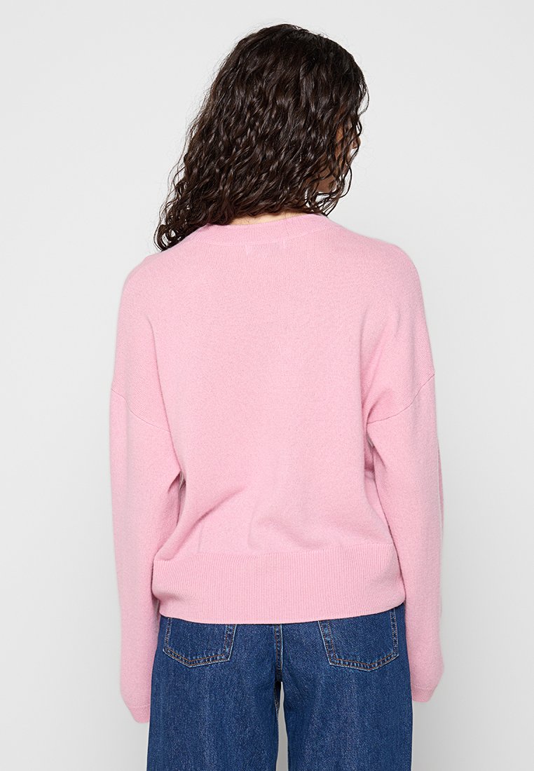 Różowy dzianinowy sweter o luźnym kroju, z opadającymi ramionami i ściągaczem na dole. Materiał wydaje się miękki, o gładkiej teksturze i minimalnych detalach.