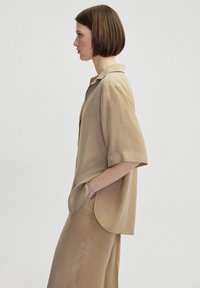 Beige, short-sleeved shirt with a pointed collar, relaxed fit, and side pockets. Smooth texture, lightweight fabric drapes gently.