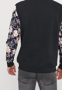 Sweat-shirt noir avec manches à motif floral, présentant des roses et des feuilles en violet, rose et vert. Matière douce avec poignets côtelés.