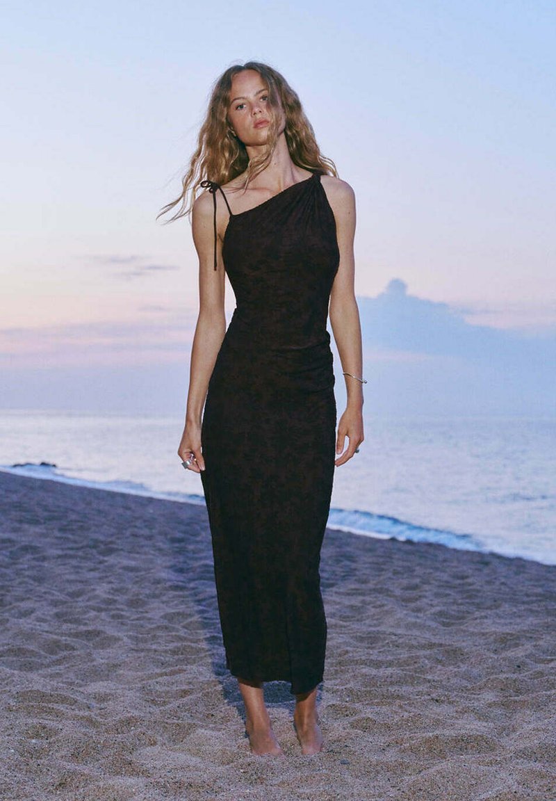 Femme aux longs cheveux portant une robe noire à une épaule, debout pieds nus sur une plage de sable au coucher du soleil au bord de la mer.