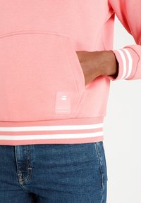 Sudadera de color rosa claro con puños a rayas blancas, dobladillo acanalado y un bolsillo frontal. Incluye una pequeña etiqueta en la esquina inferior izquierda.