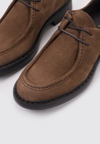 Chaussures en suède brun avec une texture lisse, bout arrondi et semelle en caoutchouc noir. Elles présentent des lacets fins et des coutures contrastantes le long des bords.