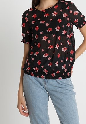 Kvinde iført en sort kortærmet bluse med rød og pink blomsterprint og rynket kant, kombineret med lyseblå jeans.