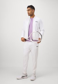 Witte sportjack met zwarte accenten, gecombineerd met een lavendelkleurig T-shirt en witte broek, met een tennisracket in de hand, staande op een effen achtergrond.