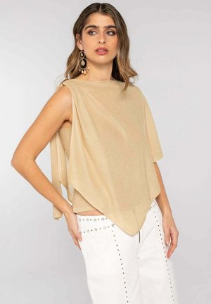 Mujer con un top beige asimétrico sin mangas combinado con pantalones blancos con tachuelas plateadas, de pie frente a un fondo liso y claro.