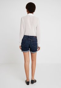 Celana pendek denim dengan ujung dilipat, warna biru tua, memiliki lima saku, dan label merek kecil di bagian belakang; dipadukan dengan blus lengan panjang putih.