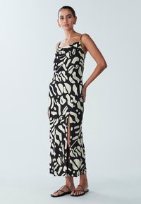 Czarna i kremowa maxi sukienka w wzory z cienkimi ramiączkami, rozcięciem z boku i drapowanym dekoltem. Noszona z czarnymi, płaskimi sandałami.
