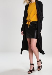 Žena ve hořčicově žluté zavázané blůze, černé kožené sukni, dlouhém černém kabátu a tmavých vysokých podpatcích, stojící proti jednoduchému pozadí.