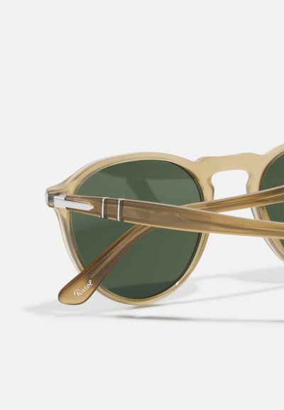 Ochelari de soare cu ramă transparentă de culoare bej deschis, lentile verzi rotunde și un accent metalic auriu pe brate, având marca "Persol."