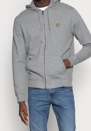 Mand iført lysegrå lynlåshætte med gul fuglelogo og blå jeans, stående med den ene hånd i lommen og den anden afslappet.