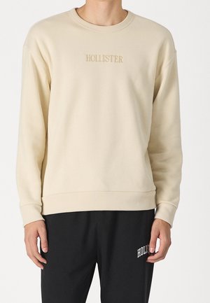 Mann trägt beige Sweatshirt mit Rundhalsausschnitt und "HOLLISTER" Text sowie schwarze Hose mit teilweisem weißen Text am linken Oberschenkel.