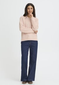 Ljusrosa stickad tröja med diamantmönster, rund halsringning och långa ärmar, matchad med vida blåjeans och bruna skor.