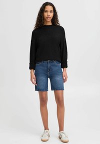 Svart texturerad tröja med vida ärmar, kombinerad med blå denimshorts. Modellen bär vita sneakers och står mot en neutral bakgrund.