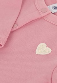 Rosa Baumwollbekleidung mit einem strukturierten, cremefarbenen Herz, das auf der Vorderseite gestickt ist. Sichtbare Knöpfe im Kragenbereich.