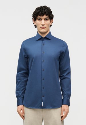 Νεαρός άνδρας με σγουρά σκουρόχρωμα μαλλιά, που φοράει εφαρμοστό, μπλε σκούρο πουκάμισο με κουμπιά και ανοιχτόχρωμο μπεζ παντελόνι, στέκεται μπροστά σε απλό φωτεινό φόντο.