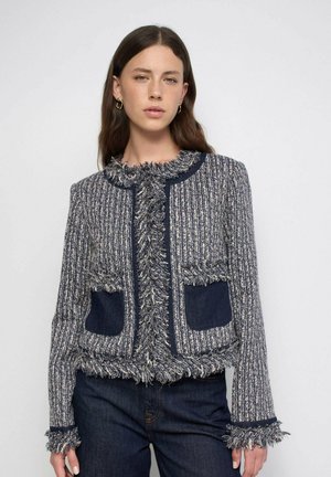 Mujer con cabello largo y castaño lleva una chaqueta gris y azul marino texturizada con bordes con flecos y vaqueros azul oscuro sobre un fondo liso.