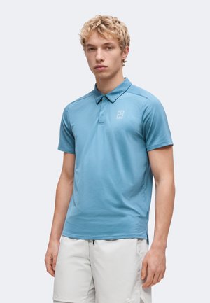 Jeune homme aux cheveux blonds bouclés, portant un polo bleu à manches courtes et un short gris clair, debout devant un fond uni.