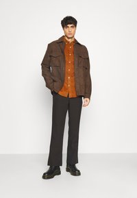 Camisa de sobrecamisa marrón con patrón de espiga y grandes bolsillos en el pecho, llevada sobre una camisa de botón de color óxido, combinada con pantalones oscuros y botas negras.