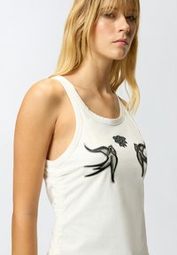 Canotta bianca con motivi di rondini ricamati neri e dettagli floreali. Tessuto morbido, design senza maniche, con arricciature laterali per aggiungere texture.