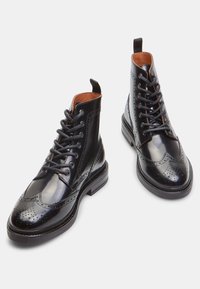 Botín de cuero negro con diseño de punta alada, perforaciones, punta redonda, cordones y tirador; presenta un acabado brillante y suela robusta.