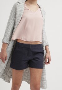 Pantalones cortos de textura azul marino con bolsillos delanteros, combinados con una blusa de manga corta rosa pálido y un abrigo de textura gris claro.