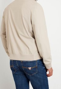 Beżowa kurtka typu bomber o gładkiej fakturze, z prążkowanymi mankietami i dołem; w połączeniu z ciemnoniebieskimi dżinsami z logo w kształcie trójkąta na tylnej kieszeni.