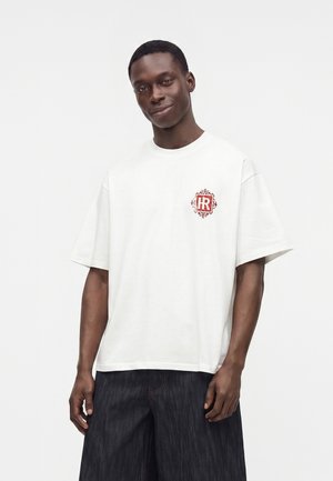 Άνδρας που φοράει λευκό υπερμεγέθες T-shirt με κόκκινο διακοσμητικό λογότυπο στο στήθος και χαλαρά σκούρα τζιν, στέκεται μπροστά σε απλό φωτεινό φόντο.