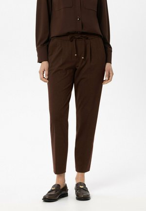 Frau trägt dunkelbraune Hose mit Kordelzug, passendes langärmeliges Shirt und dunkelbraune Loafer, sie steht auf weißem Hintergrund.