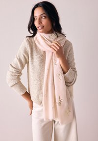 Scarf rosa chiaro con ricami floreali e accenti verdi. Tessuto morbido e bordi frangiati. Indossato con un maglione beige sopra pantaloni bianchi.
