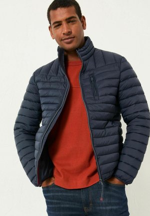 Navy puffer jacket with vertical stitching, high collar, and zippered pocket, worn over a rust-colored shirt and jeans.
