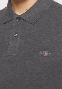 Polo gris en tissu doux, avec col pointu, deux boutons, et logo brodé avec un motif de blason sur la poitrine.