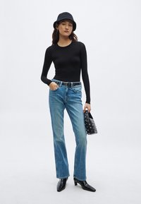 Zwart top met lange mouwen, lichtblauwe jeans met rechte pijpen, zwarte gestructureerde handtas met studs, en zwarte enkellaarzen met hak. Neutrale achtergrond.