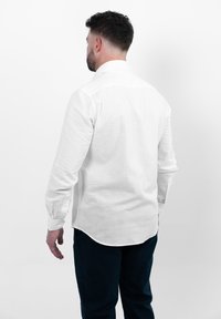 Witte linnen overhemd met lange mouwen en een gebogen zoom, gecombineerd met donkerblauwe broeken. Het achteraanzicht toont een gladde textuur en eenvoudig ontwerp.