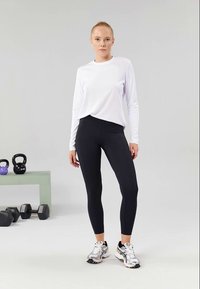 Fehér, hosszúujjú sportfelső fekete leggingsszel párosítva, színes sportcipőkkel kiegészítve. A háttérben edzőfelszerelések láthatók.