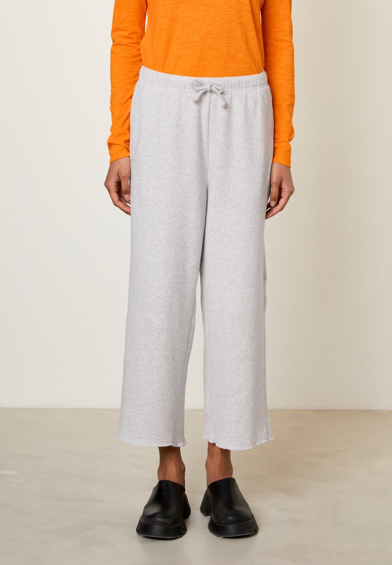 Lichtgrijze sweatpants met een taillekoortje, wijd uitlopende pijpen en een onafgewerkte zoom. Gecombineerd met een oranje top met lange mouwen en zwarte instapschoenen.